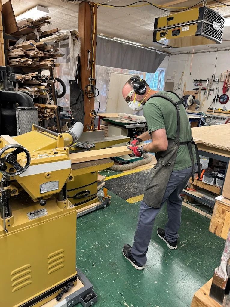 A woodworker wearing safety gear feeds a wooden board through a yellow jointer machine in a workshop.