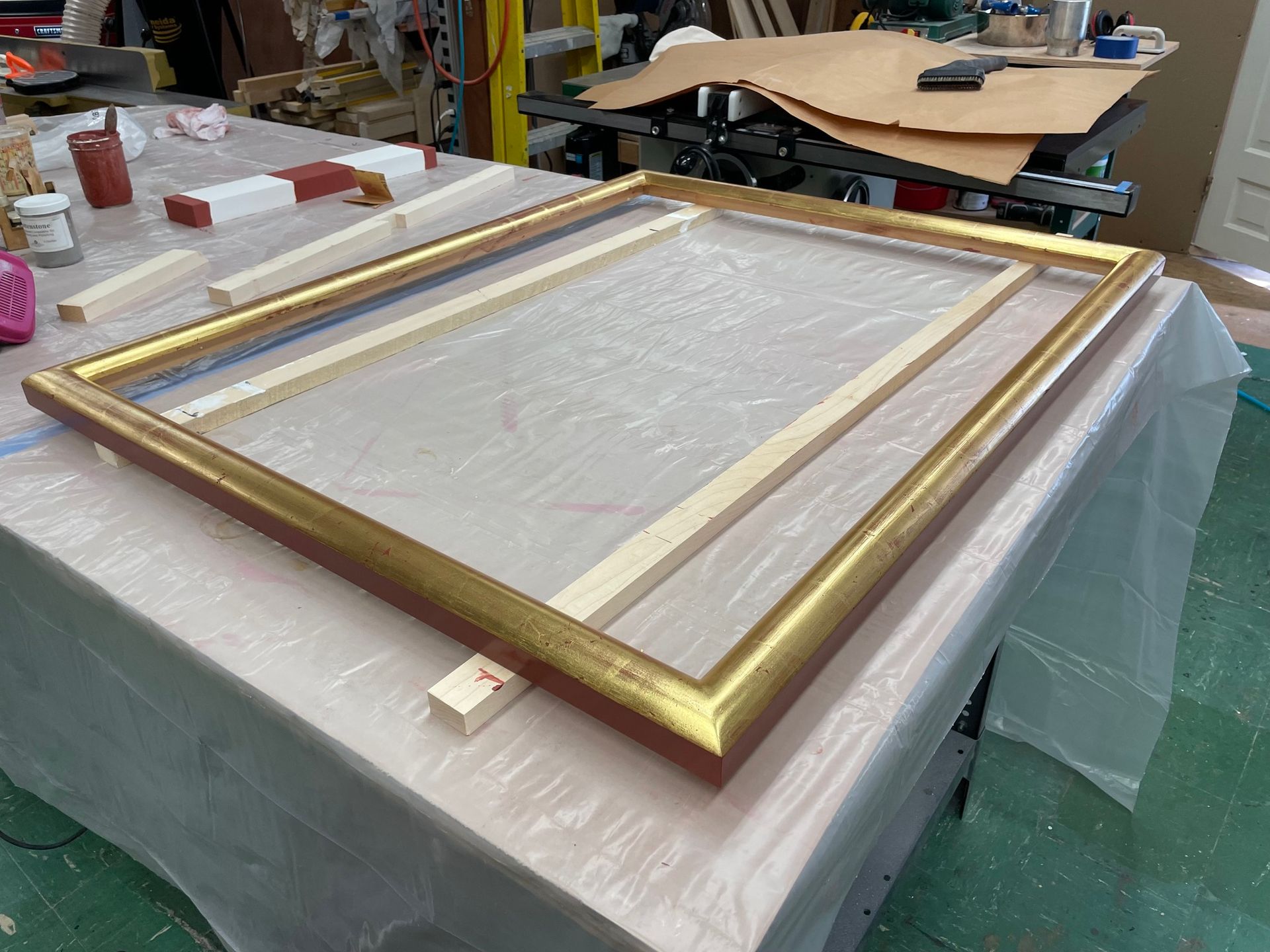 An ornate gold-leaf picture frame lies flat on a plastic-covered workbench in a workshop.