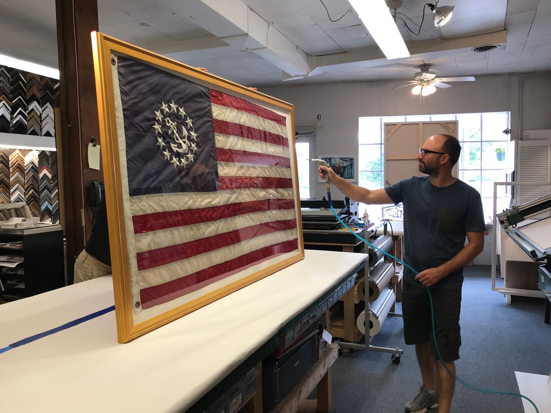 A person uses a spray tool to work on a large, framed American flag with an anchor emblem in a workshop setting.