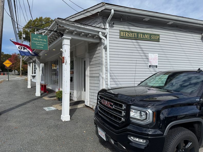 A black pickup truck parked in front of a white, single-story storefront building with a green sign reading 'HERRLY PANO'.