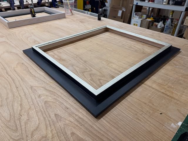 A rectangular silver metal frame mounted on a flat black backing board, sitting on a wooden workbench in a workshop.