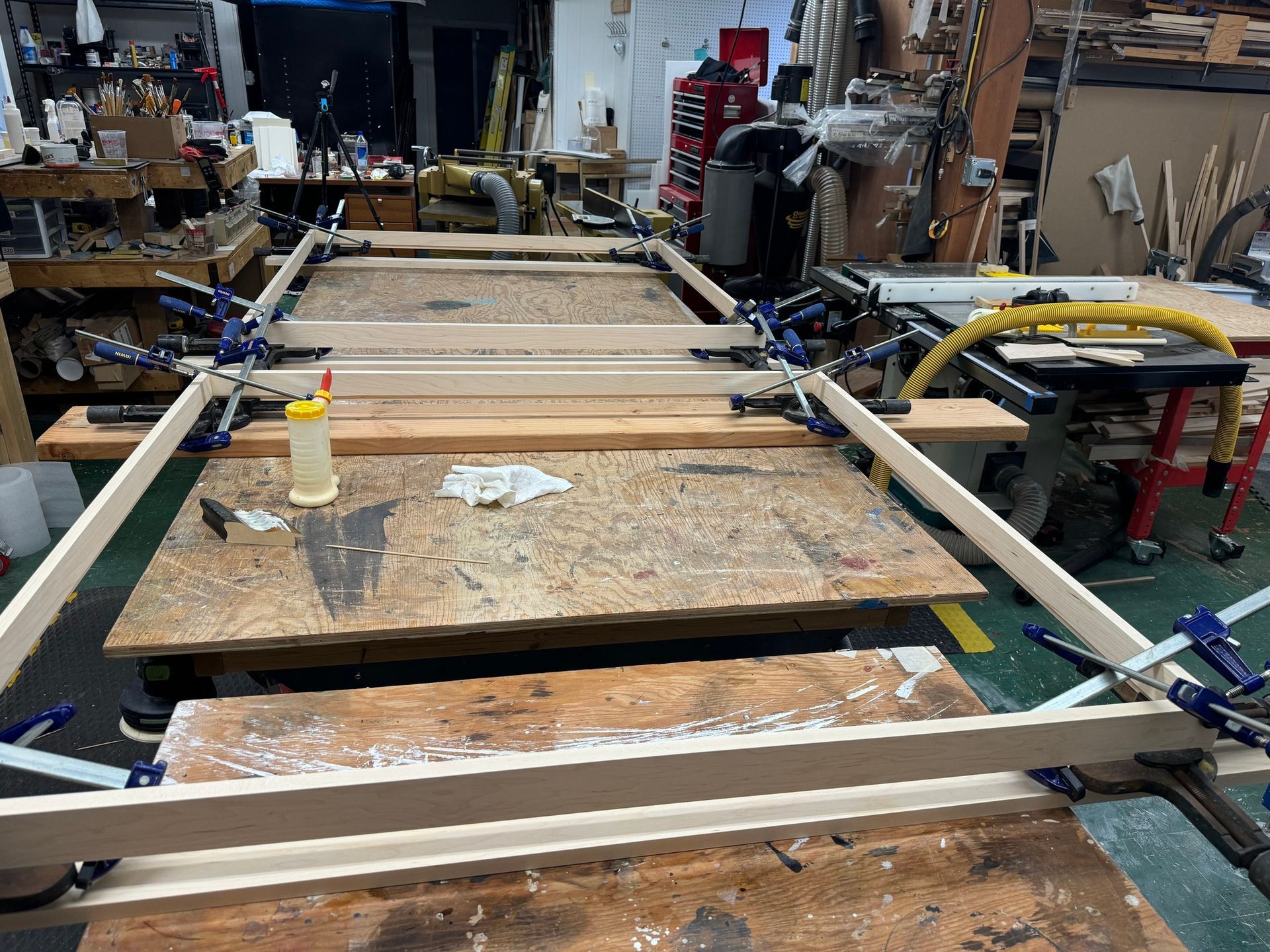 A wooden frame is being glued and clamped together on a workbench in a cluttered woodshop.