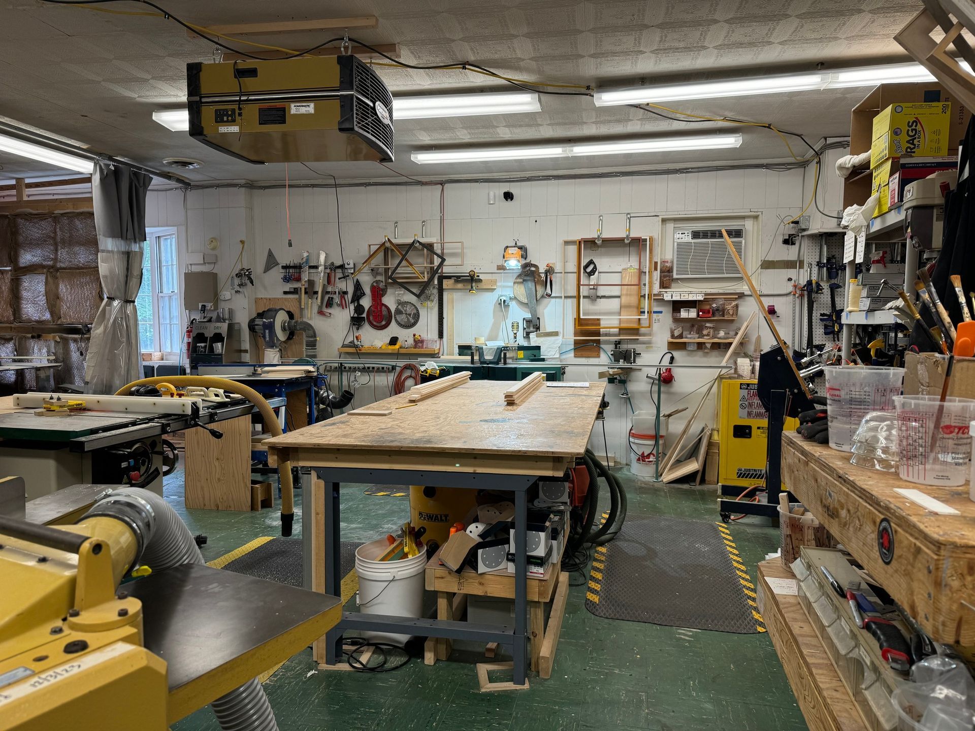 A well-equipped woodworking workshop with a large central workbench, yellow machinery, and various tools on the walls.