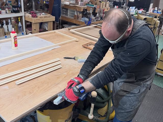 A person wearing safety glasses and red work gloves sands a piece of wood on a workbench in a woodworking shop.