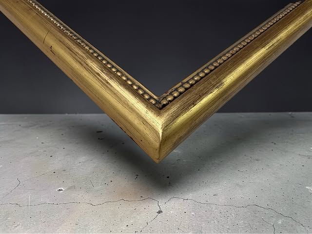 A decorative gold picture frame corner angled against a dark background, resting on a gray concrete surface.