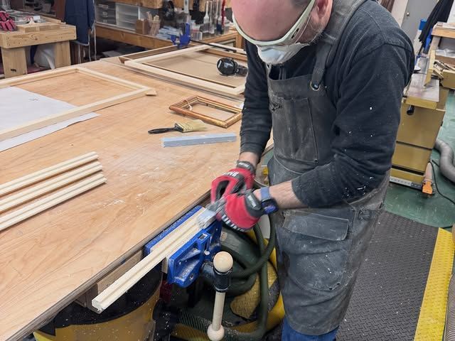 A woodworker wears a mask and safety goggles while using a hand tool to shape a piece of wood held in a workbench vise.