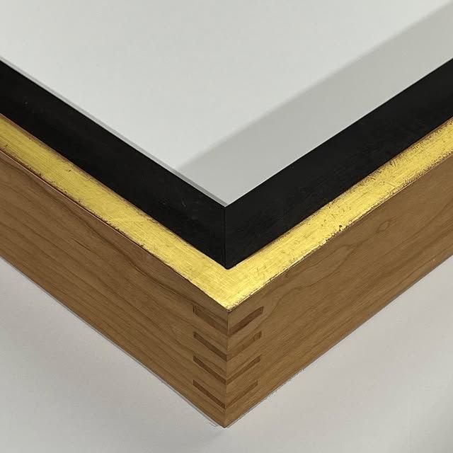 A close-up of a wooden picture frame corner with visible box joints, a gold inner rim, and a black accent border.