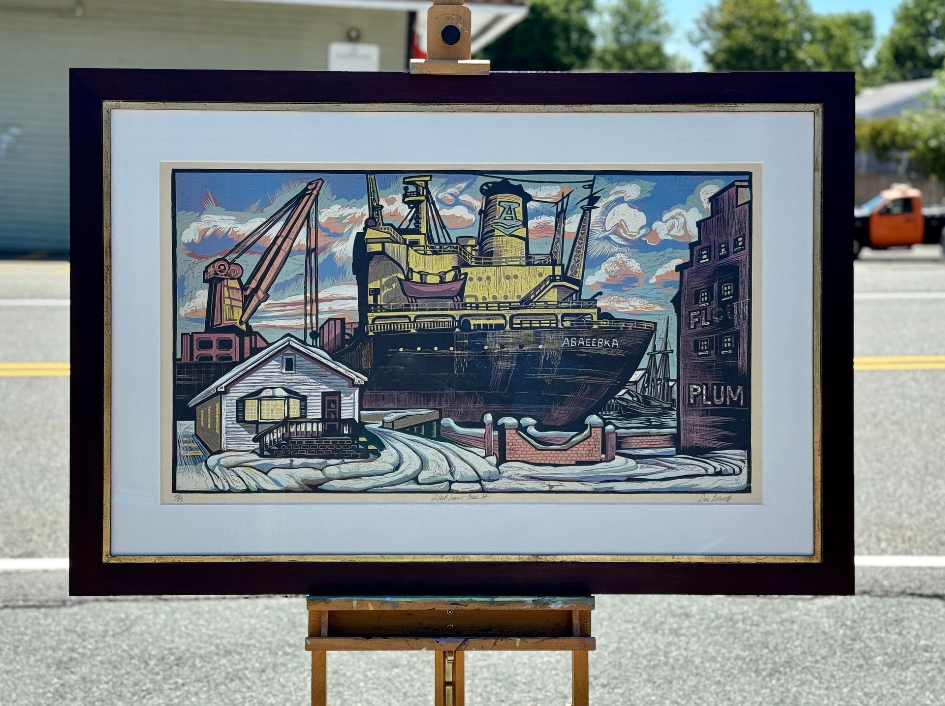 A framed woodcut print showing a large yellow and black ship docked at an industrial harbor with a crane and building.