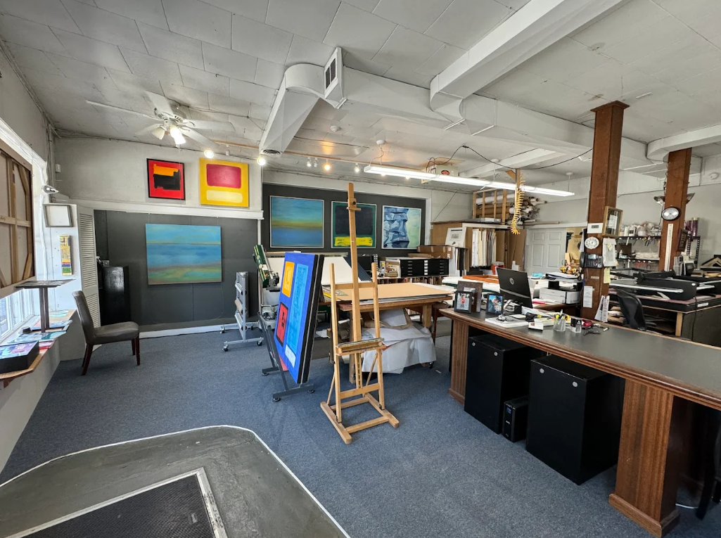 A person wearing safety glasses and red work gloves sands a piece of wood on a workbench in a woodworking shop.