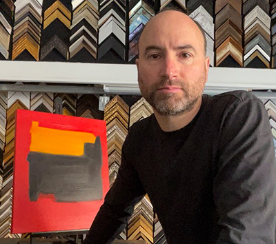 A person in a dark long-sleeved shirt stands before a framed abstract painting with orange and grey shapes on a red field.