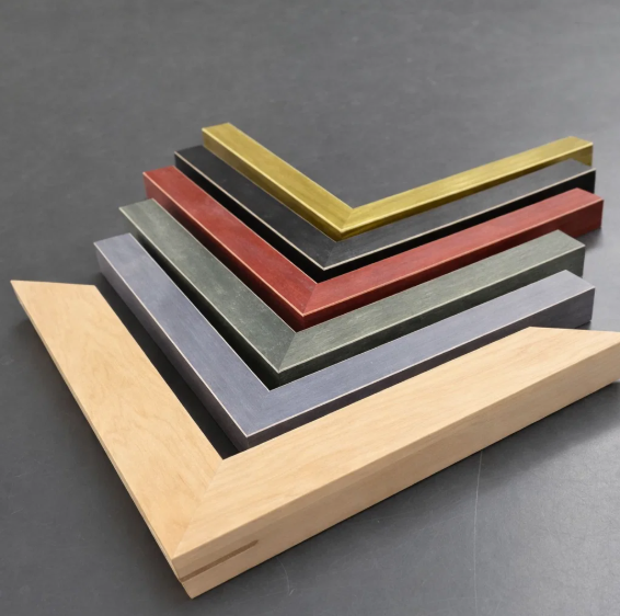 A close-up of a wooden picture frame corner with visible box joints, a gold inner rim, and a black accent border.