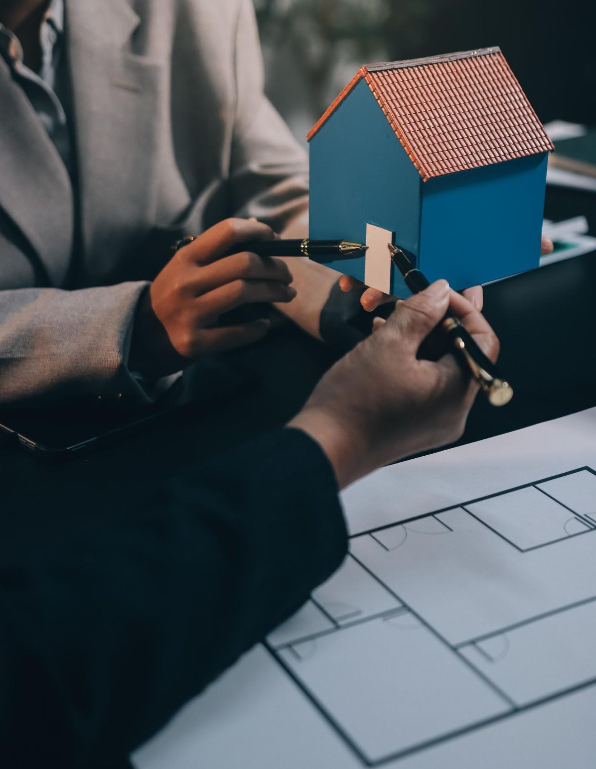 Two people examine a miniature blue house and floor plan, pointing with pens.