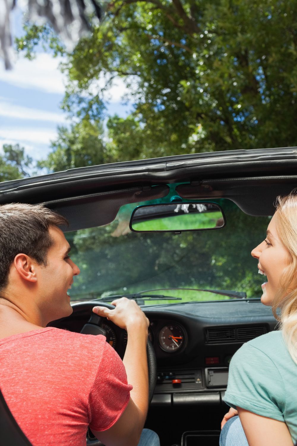Couple in a convertible car, smiling while driving through a green, wooded area on a sunny day.