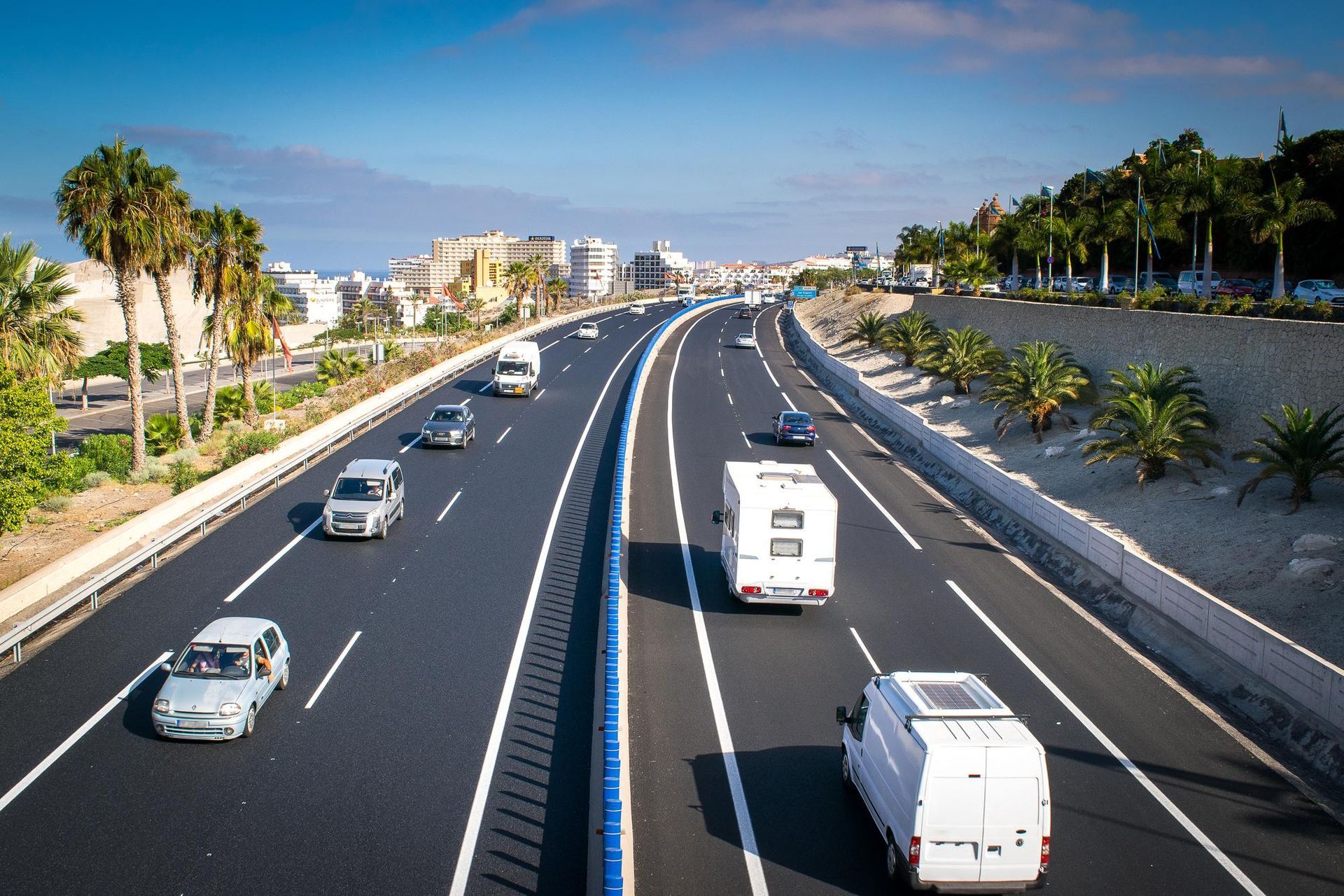 Cars and vans driving on a multi-lane highway, separated by a blue barrier, under a blue sky, palm trees alongside.