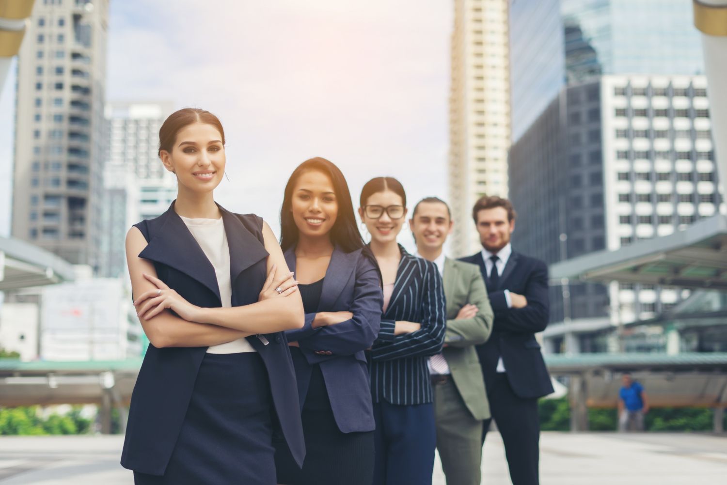 Business team standing, arms crossed, smiling. Skyscrapers in background. Sunny outdoor setting.