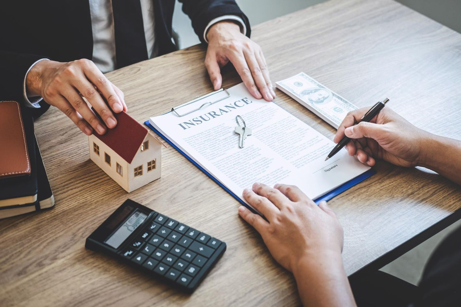 Person signing insurance document, toy house, calculator, cash on desk.