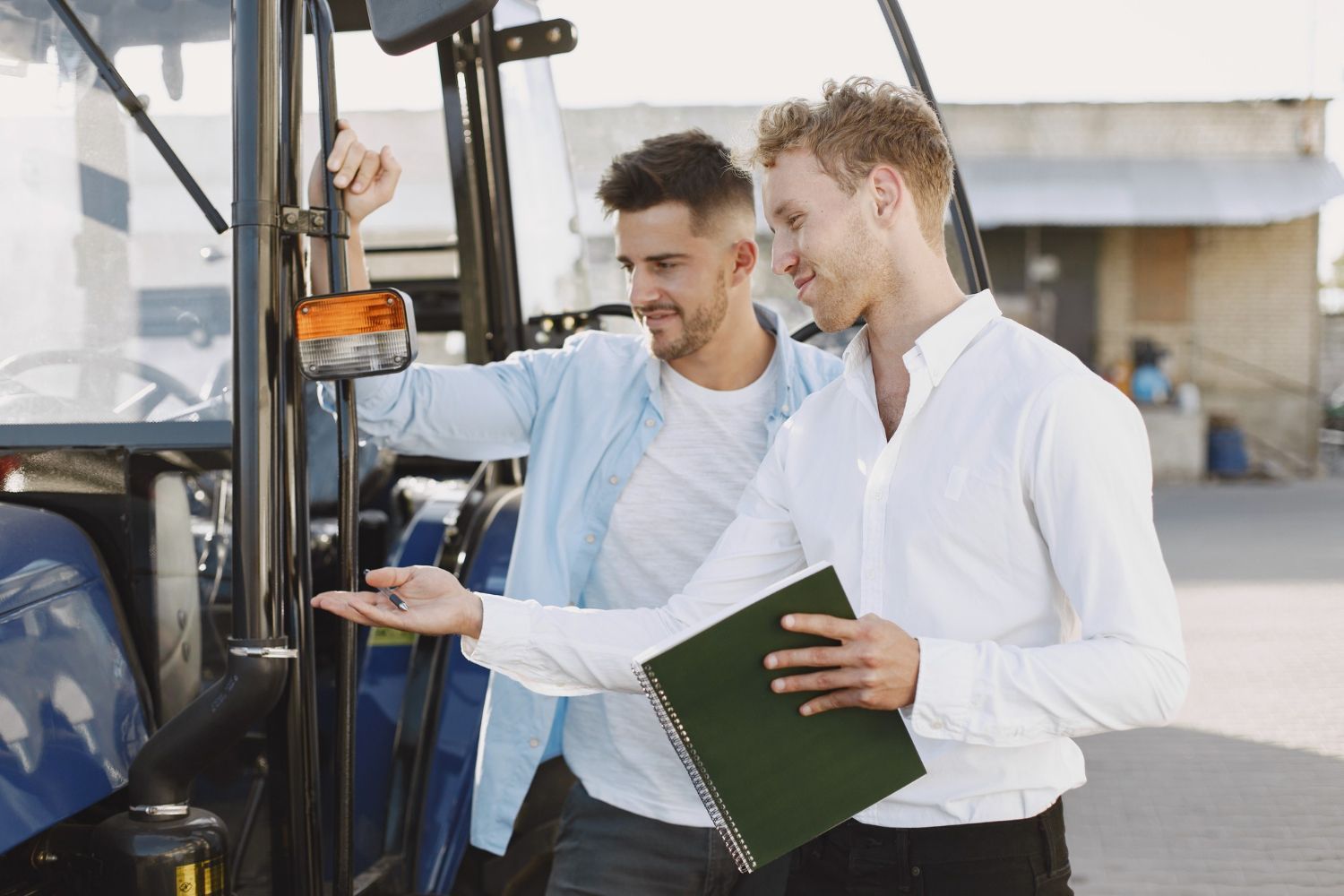 Two men examining a tractor. One points, the other holds a notebook. Outdoors, sunny.