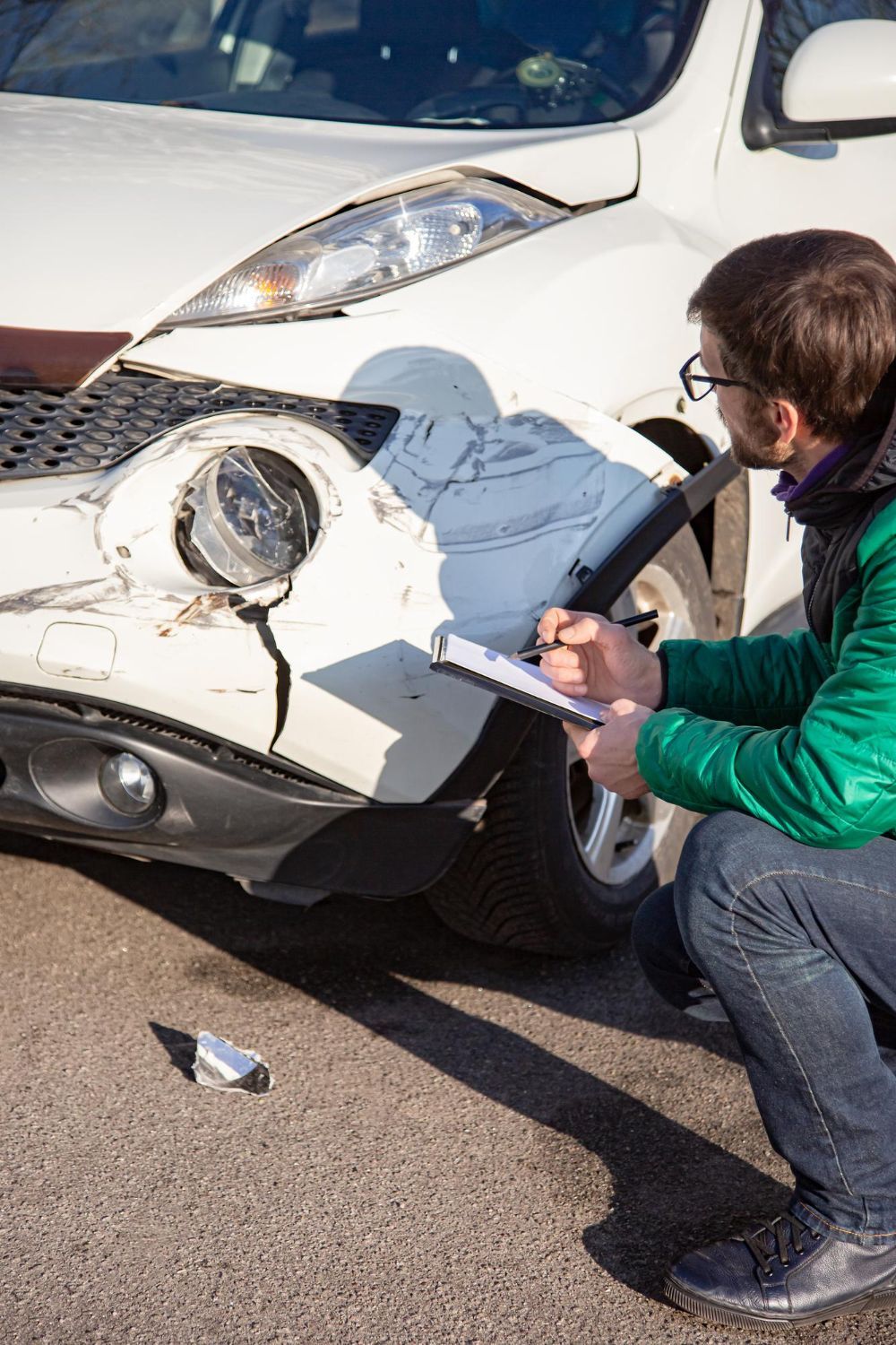 Man inspecting damage to a white car after a collision, writing on a notepad.