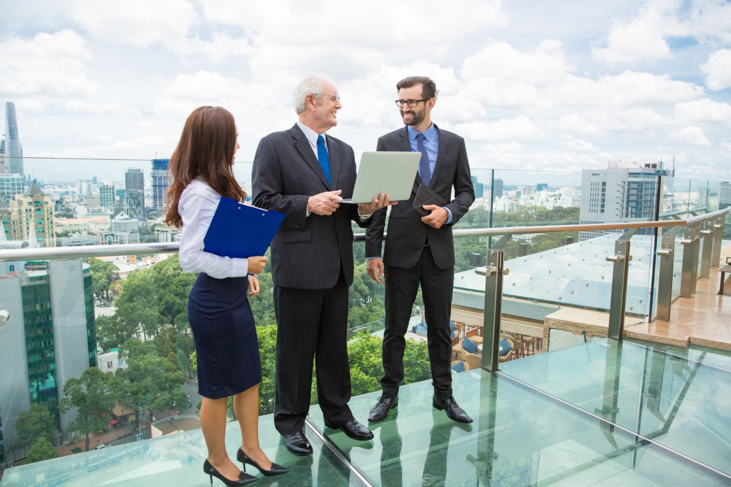 Three businesspeople on a rooftop, looking at a laptop. City view in background.