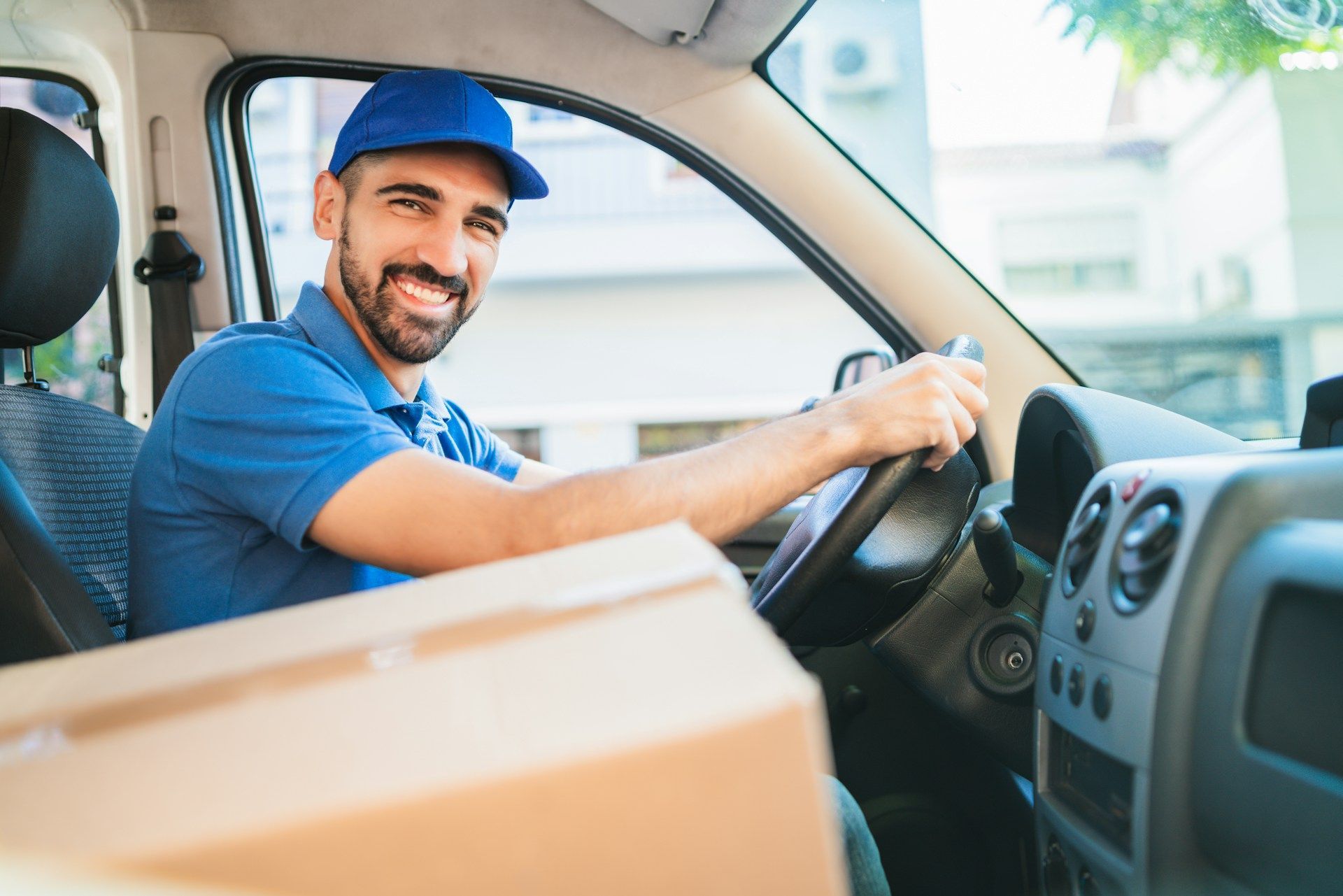 Delivery driver smiling in a van, holding a package, wearing blue shirt and cap.