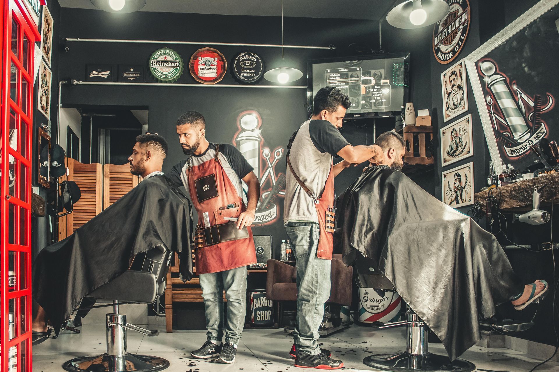 Two barbers cutting hair in a shop; black aprons, dark walls with decor, and a red phone booth structure on left.