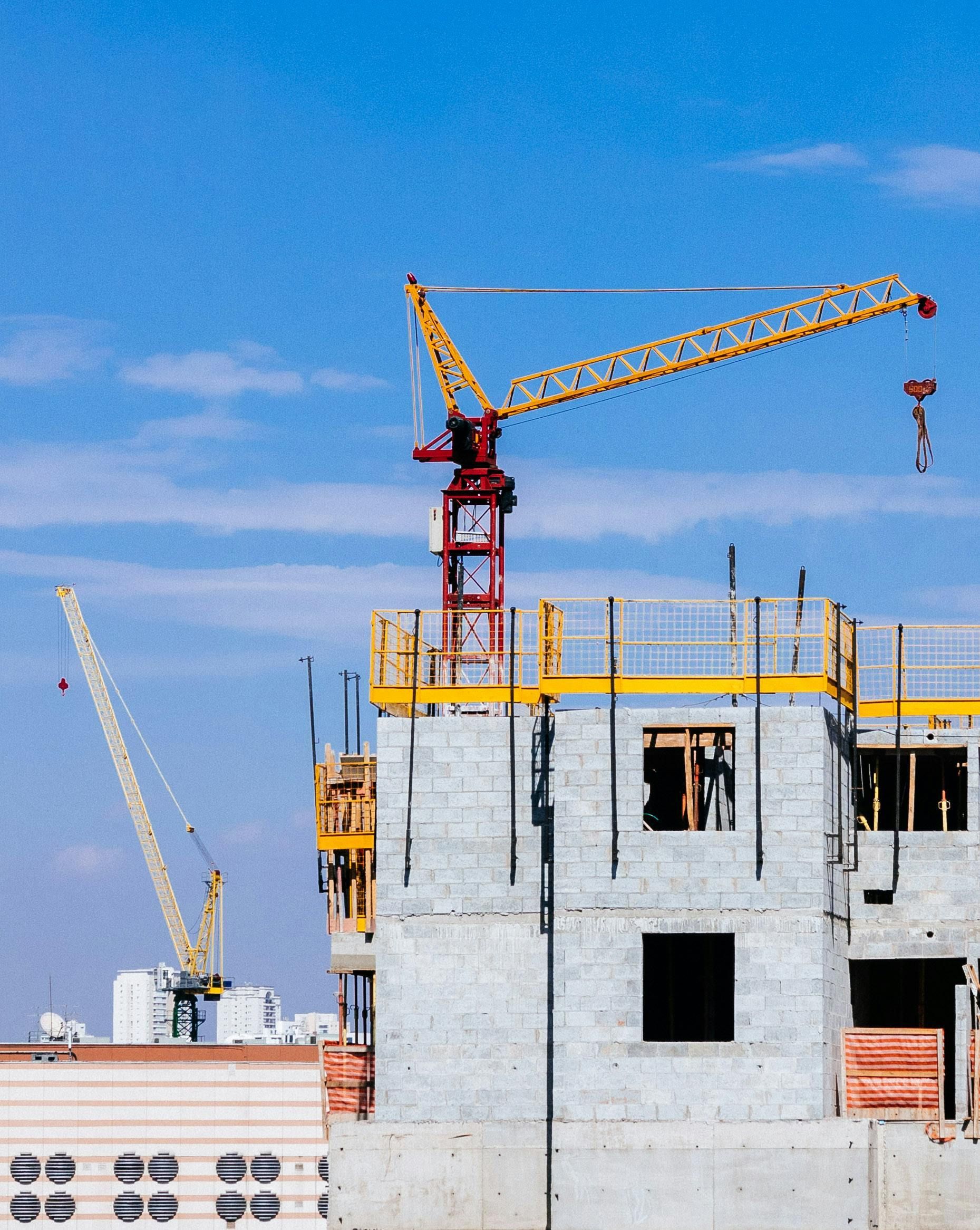 Construction site with two cranes lifting materials. Concrete building in progress against a blue sky.
