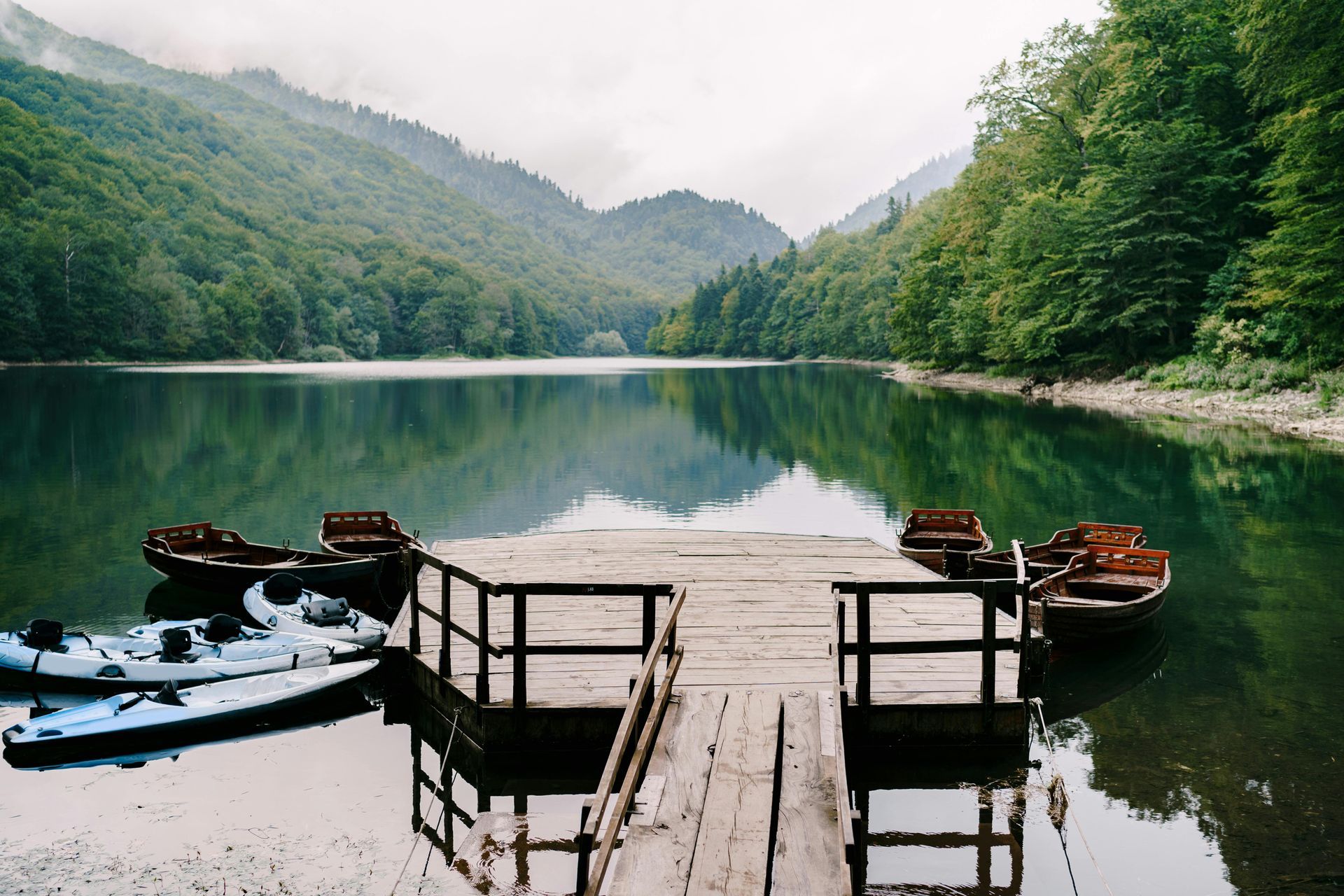 Wooden dock with kayaks and boats on a calm lake, surrounded by mountains and trees.