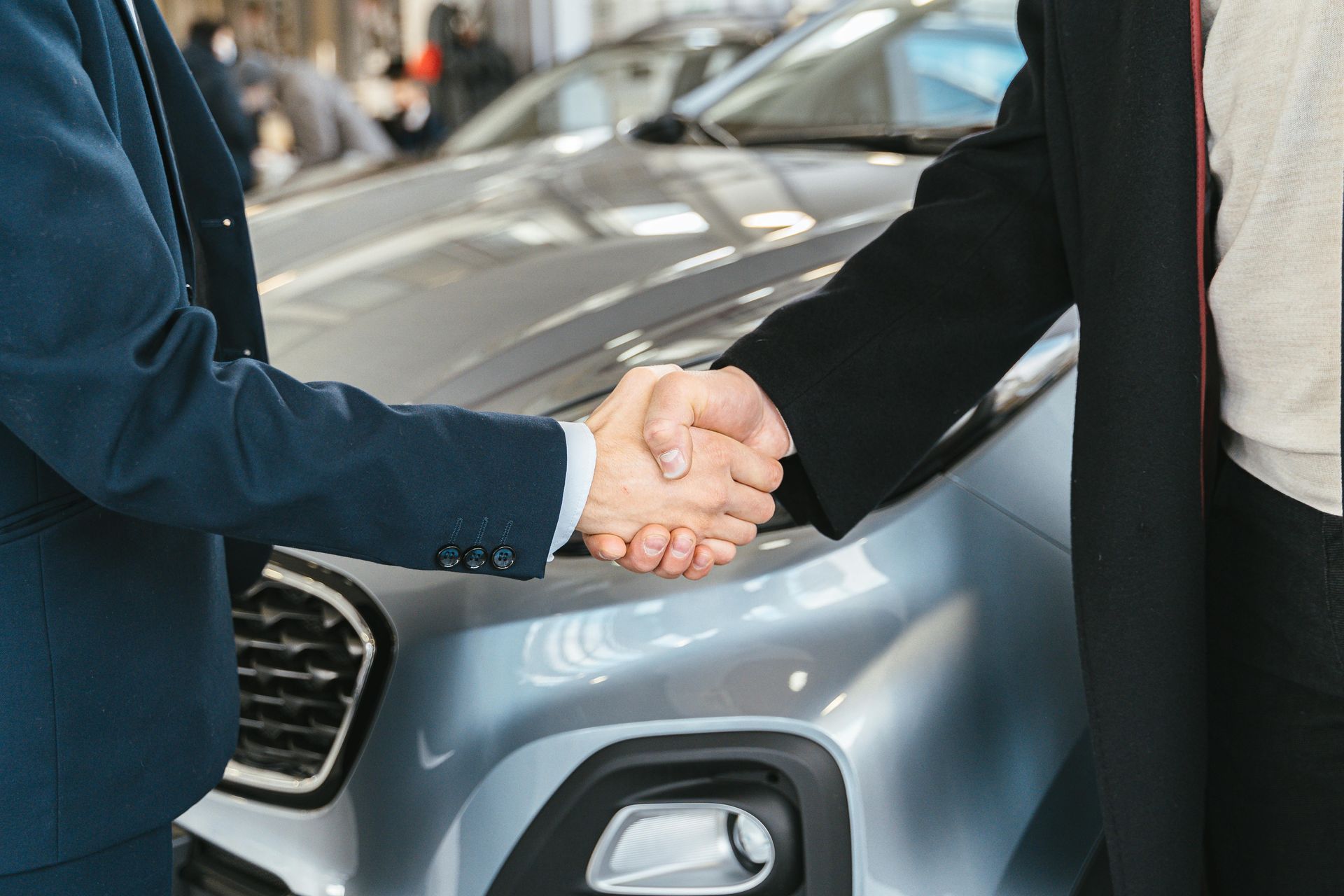 Two people shaking hands in front of a silver car in a dealership.