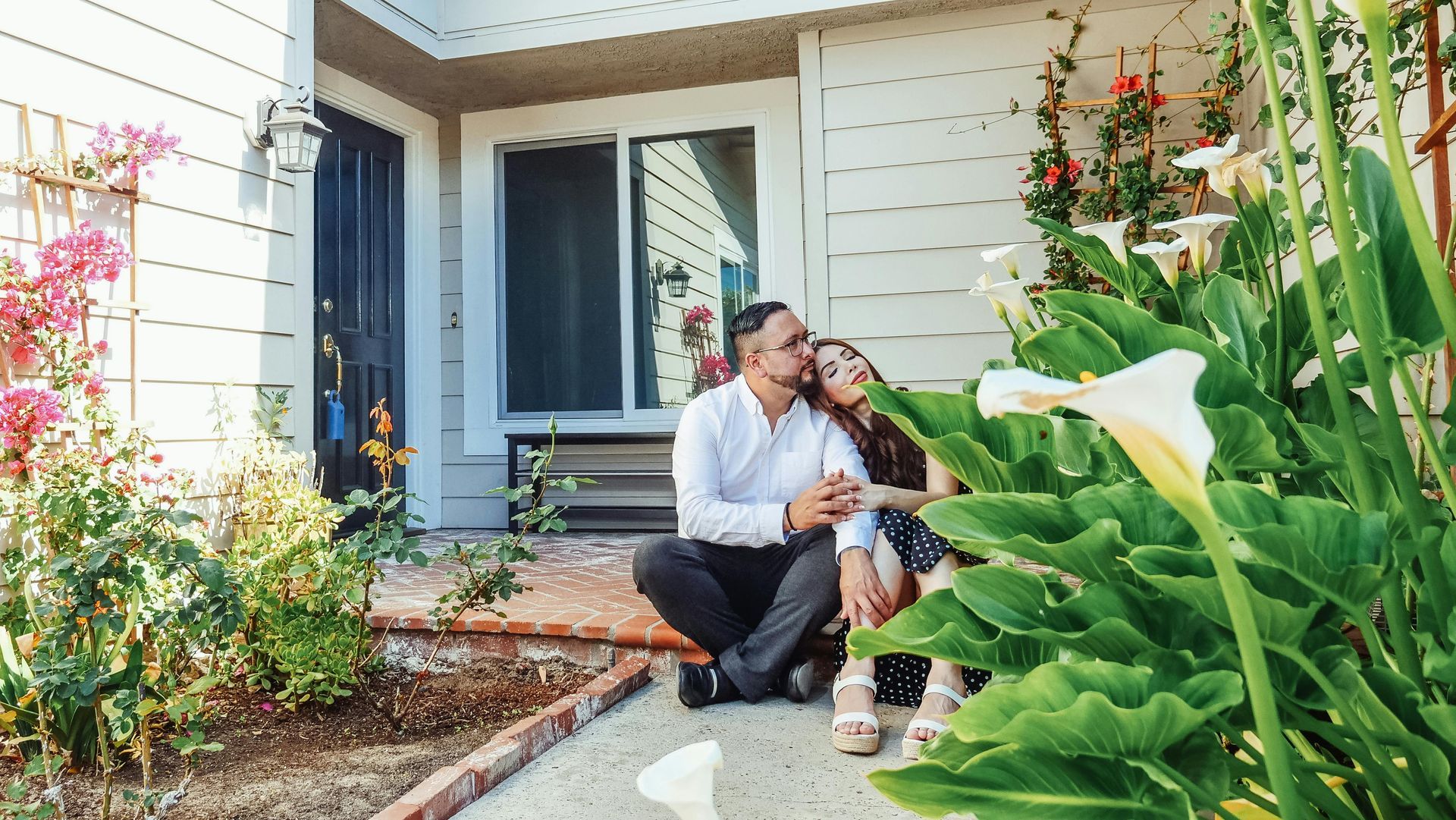 Couple sitting on a porch, embracing near flowers and a home entrance.