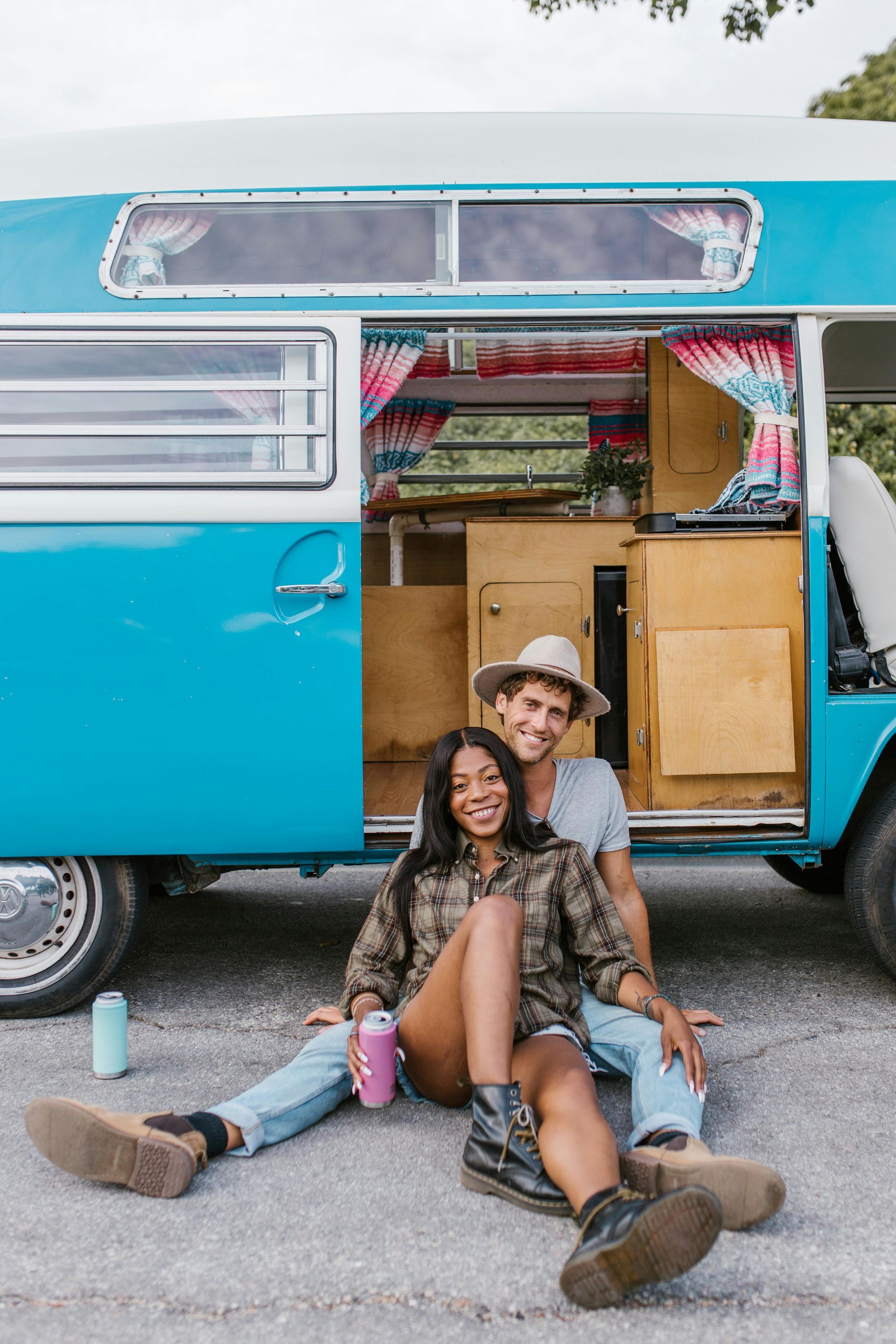 Couple sits in front of blue camper van. Smiling, woman holds drink, man wears hat.