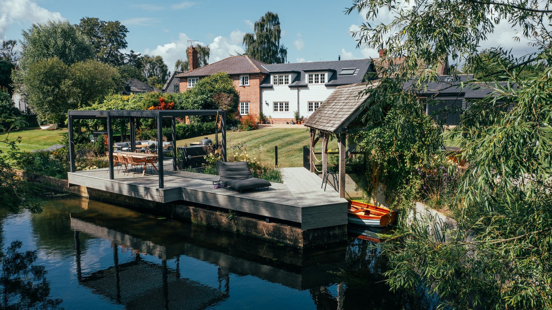 House with dock on water, outdoor seating, trees, kayaks. Bright sunny day, blue sky, reflection in water.