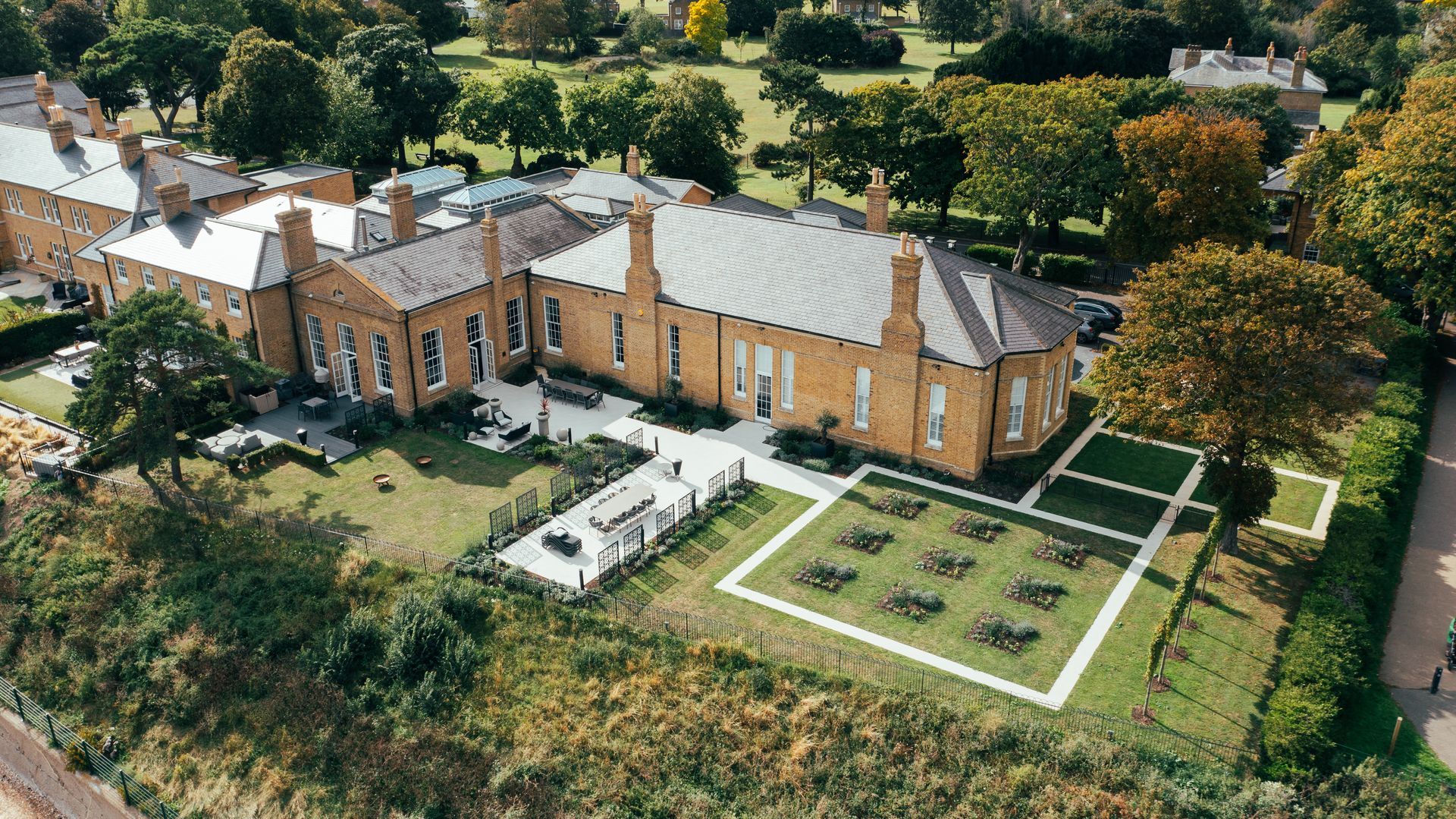 Aerial view of a long brick building with a gray roof and surrounding greenery; outdoor seating.