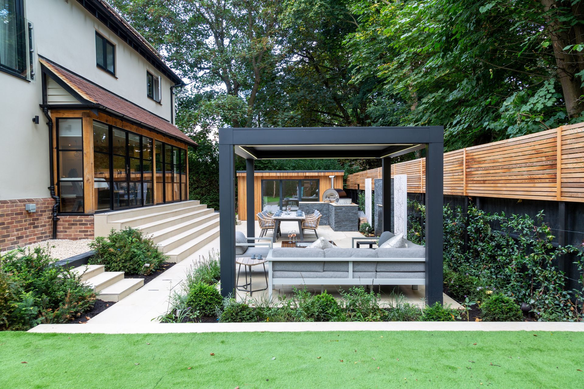 Outdoor patio with modern pergola, concrete furniture, and a house in the background. Green lawn and steps.
