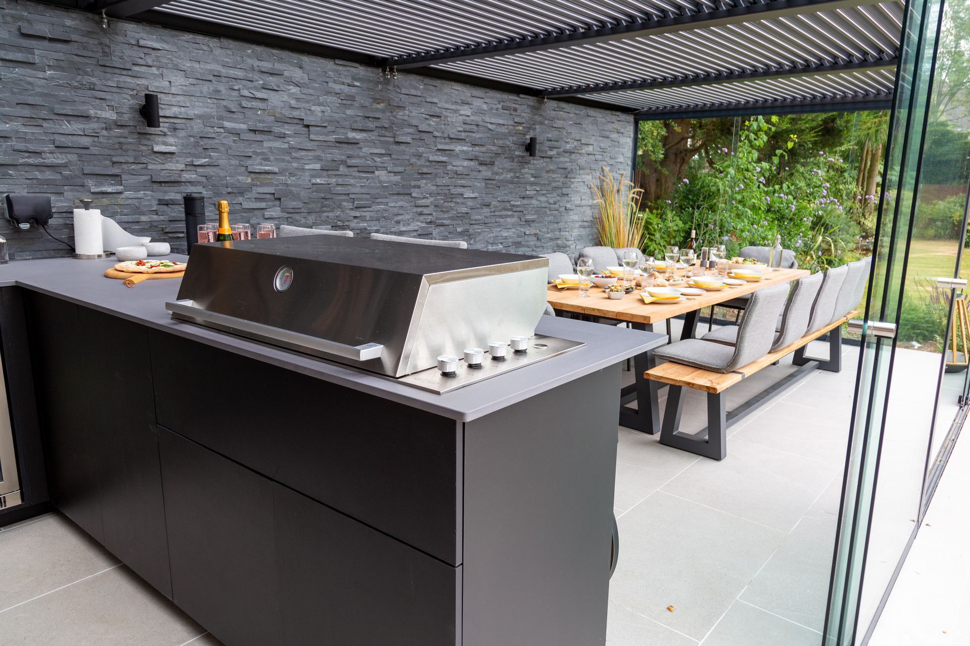 Outdoor kitchen with grill, dining table, and stone wall under a pergola.