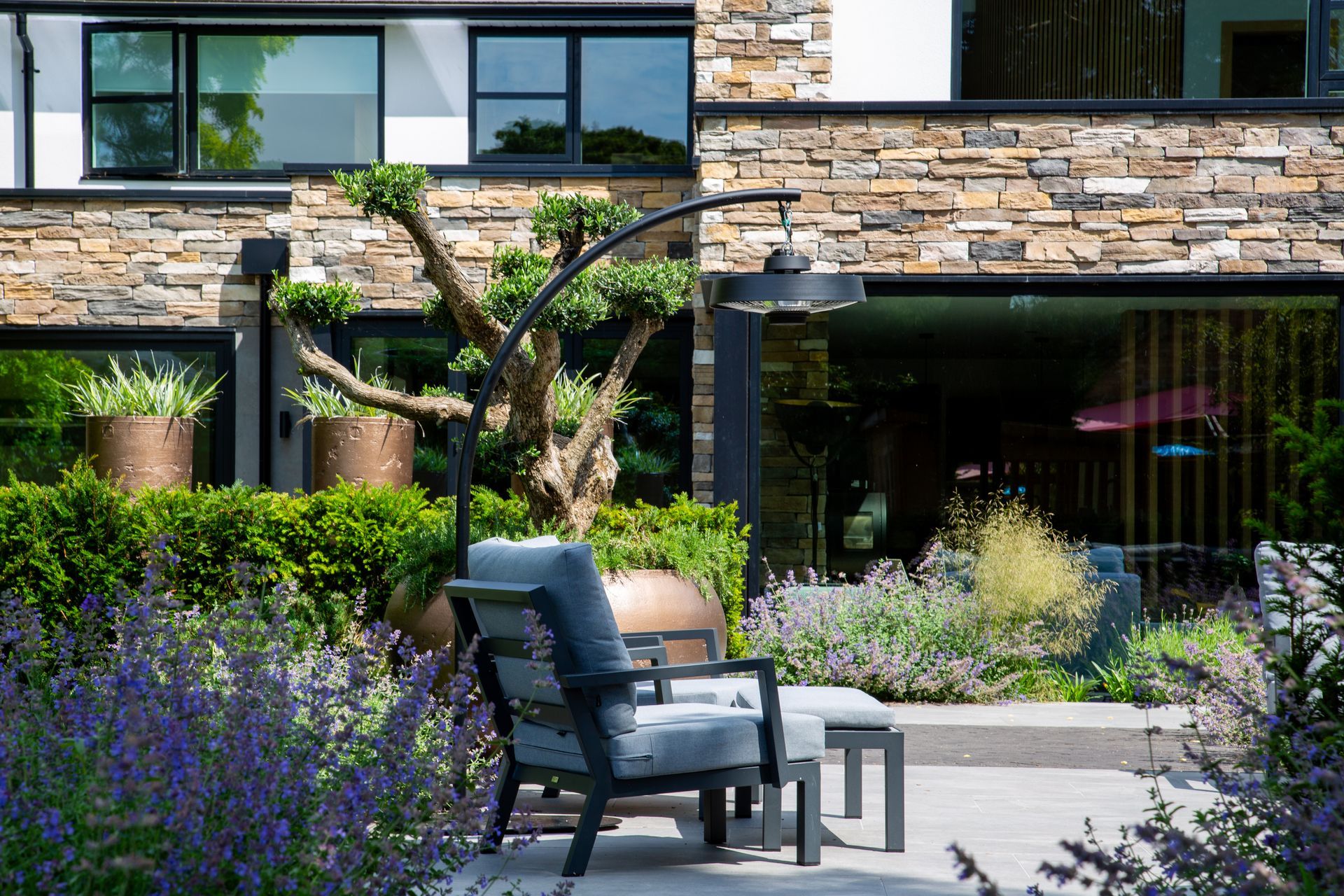 Patio with chair, ottoman, and heat lamp in front of a modern home, surrounded by plants and garden beds.