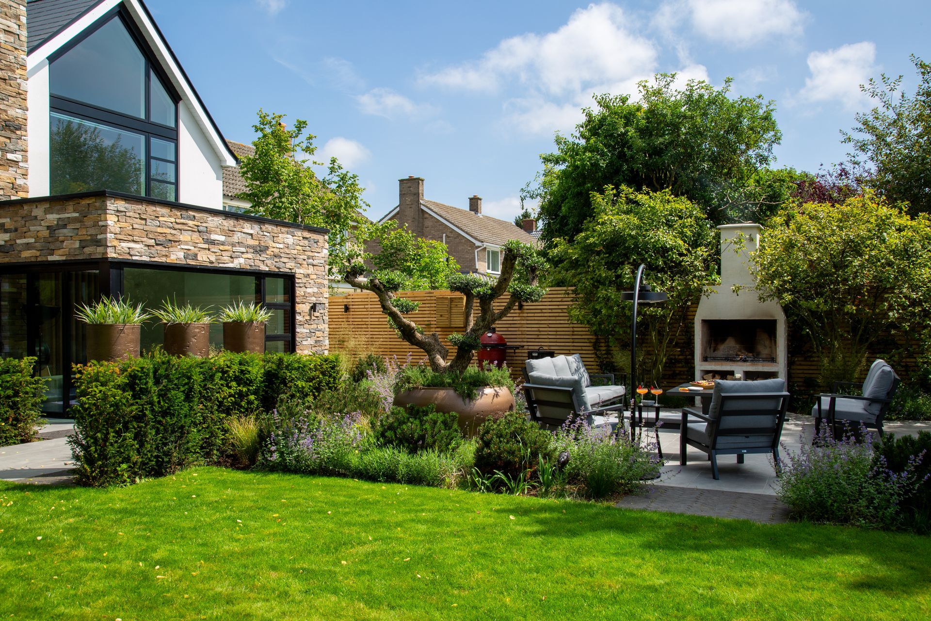Backyard patio with seating, fireplace, and manicured landscaping in front of a modern home with a stone facade.