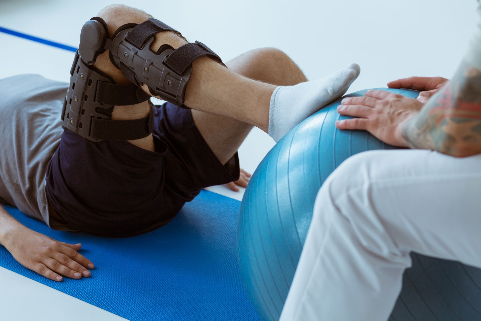 Person in knee brace doing physical therapy, foot on blue exercise ball, assisted by therapist.