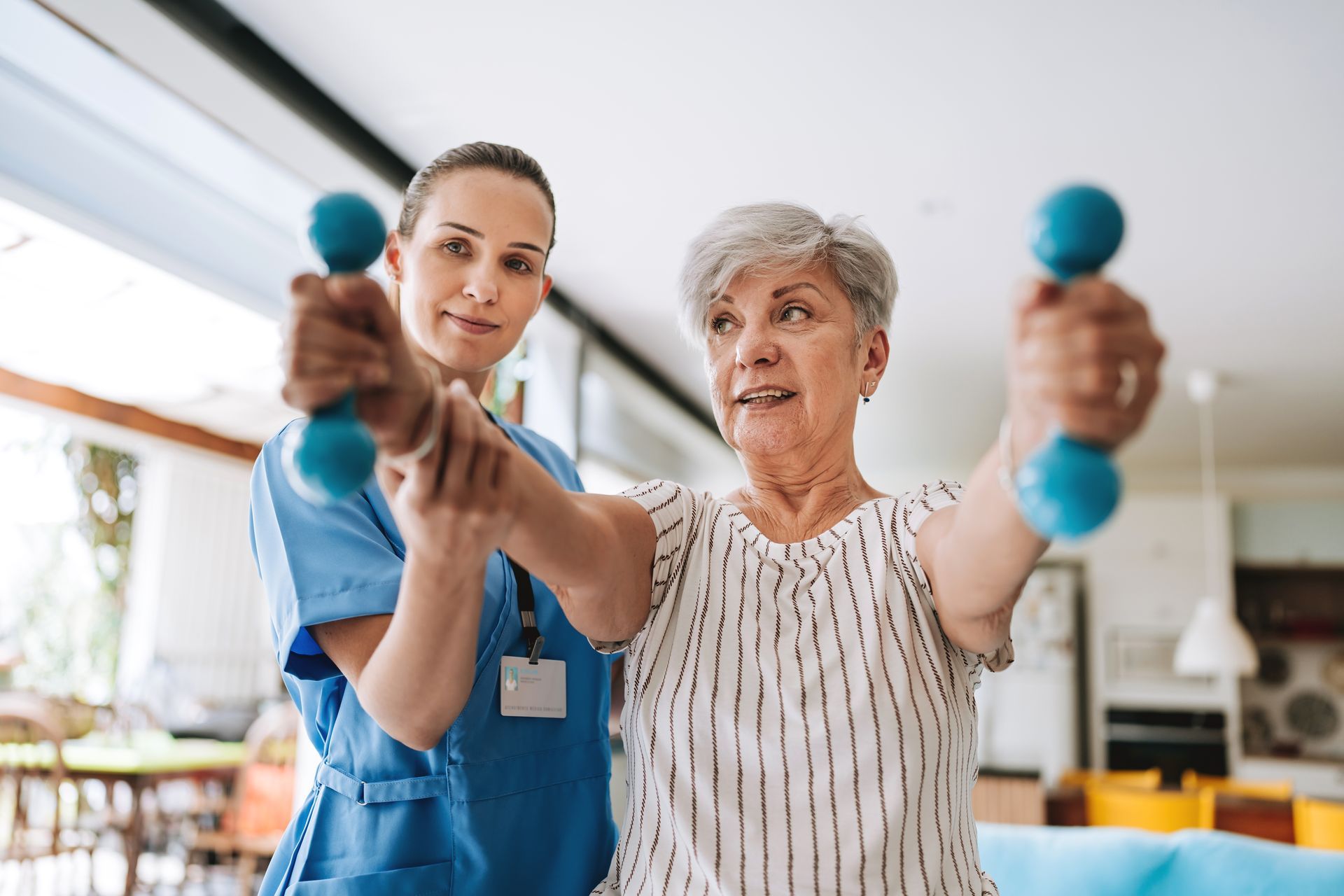 A healthcare worker assists an elderly woman with dumbbell exercises in a home setting.