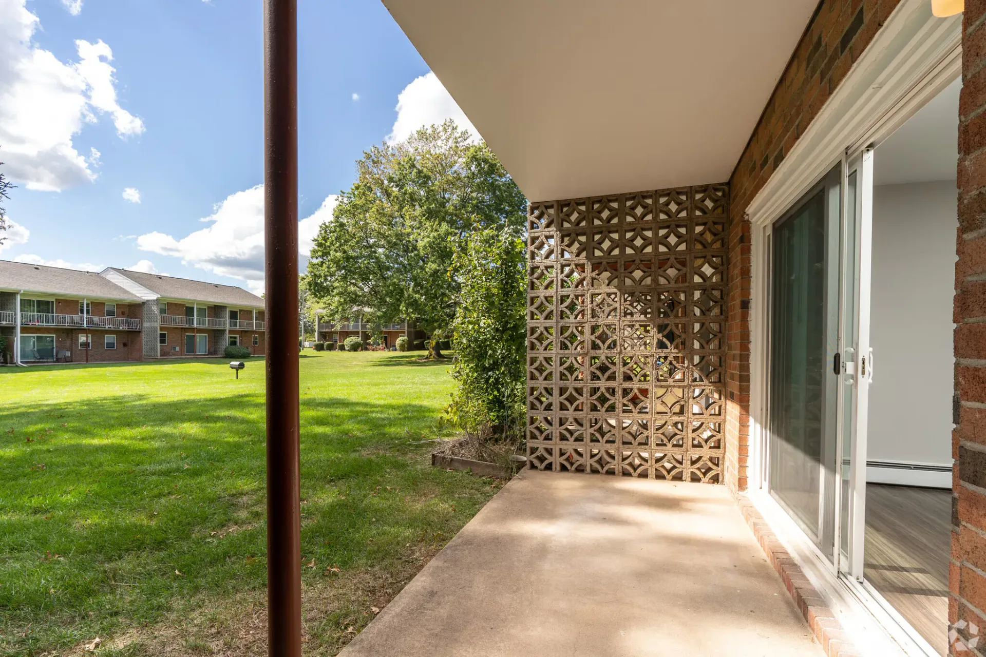 Ground-level patio with a sliding glass door, brick wall, decorative lattice, and a grassy apartment courtyard.