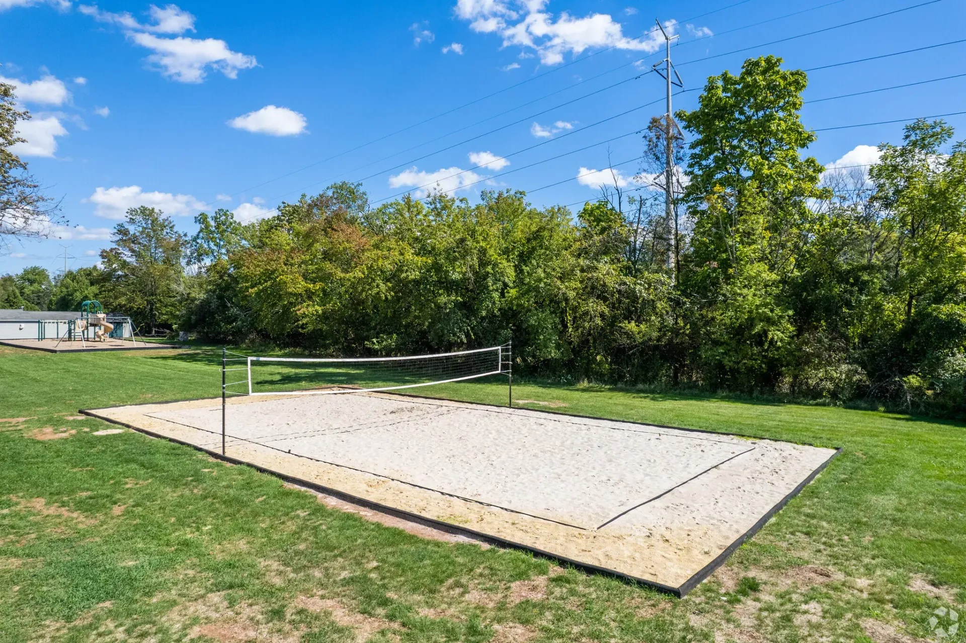 Sand volleyball court on a grassy field with a net, trees, and a playground in the background.
