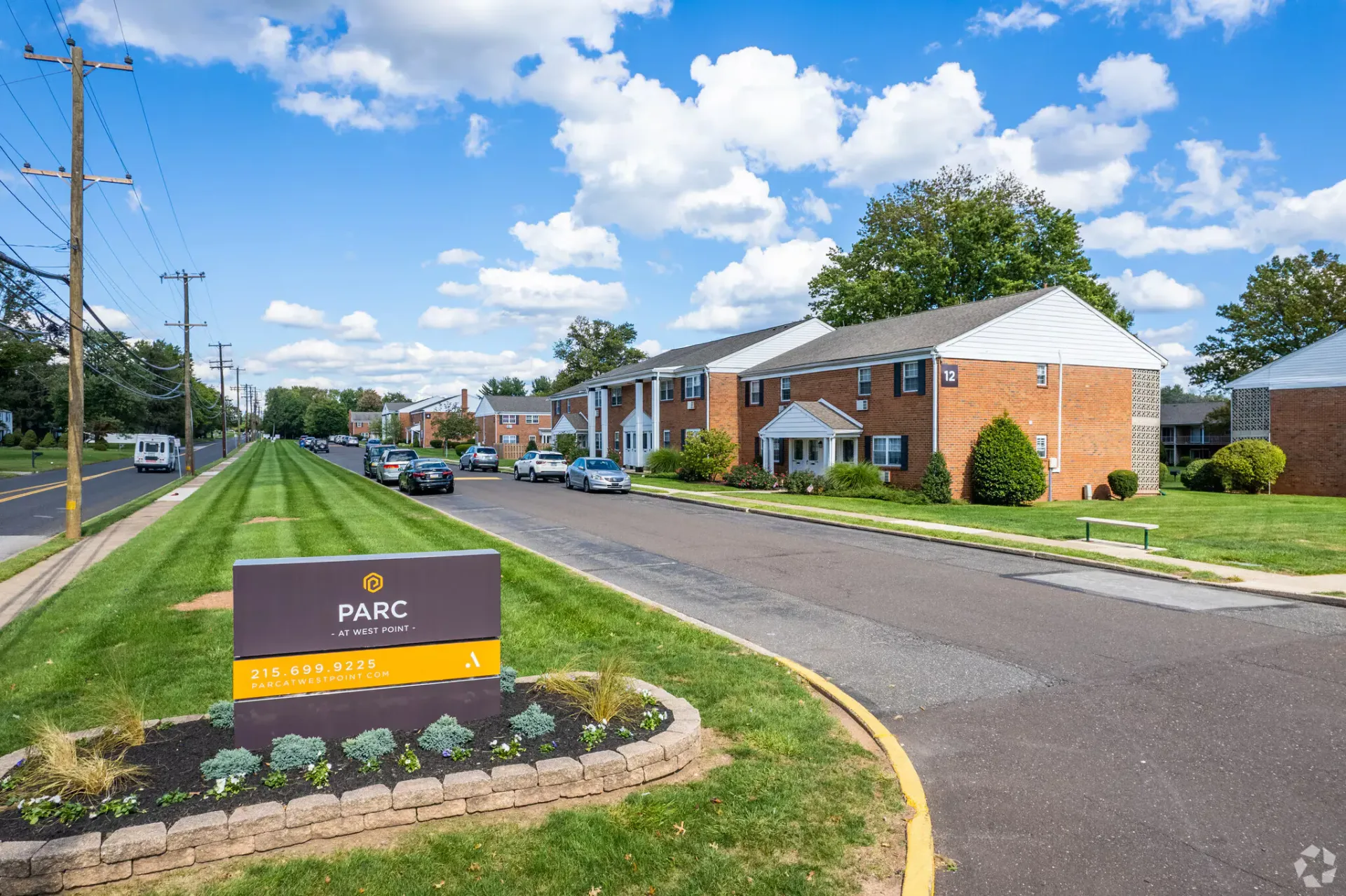 Exterior view of a brick apartment community along a tree-lined street with a prominent sign in the foreground.