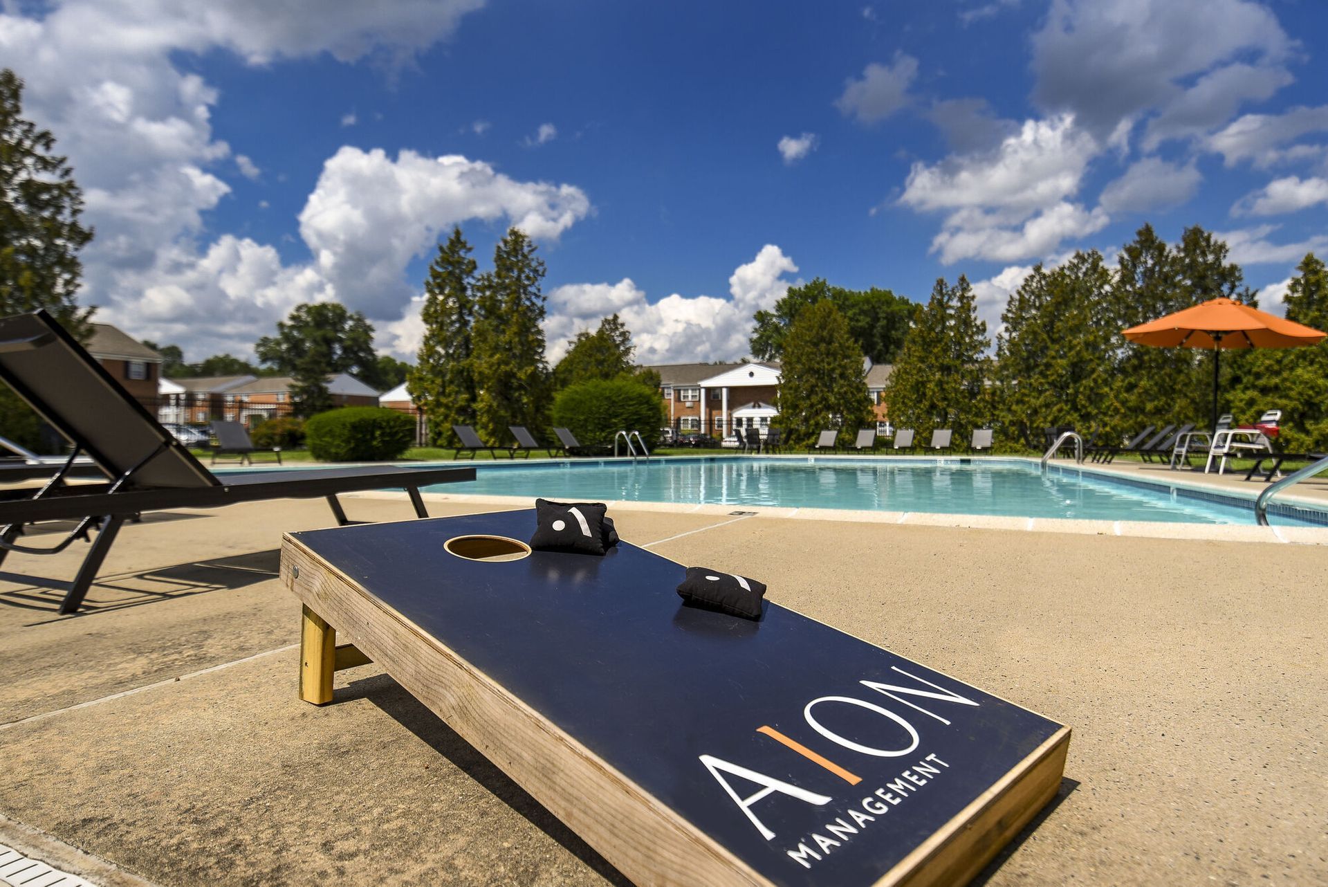 Outdoor apartment community pool area with lounge chairs and a foreground cornhole board.