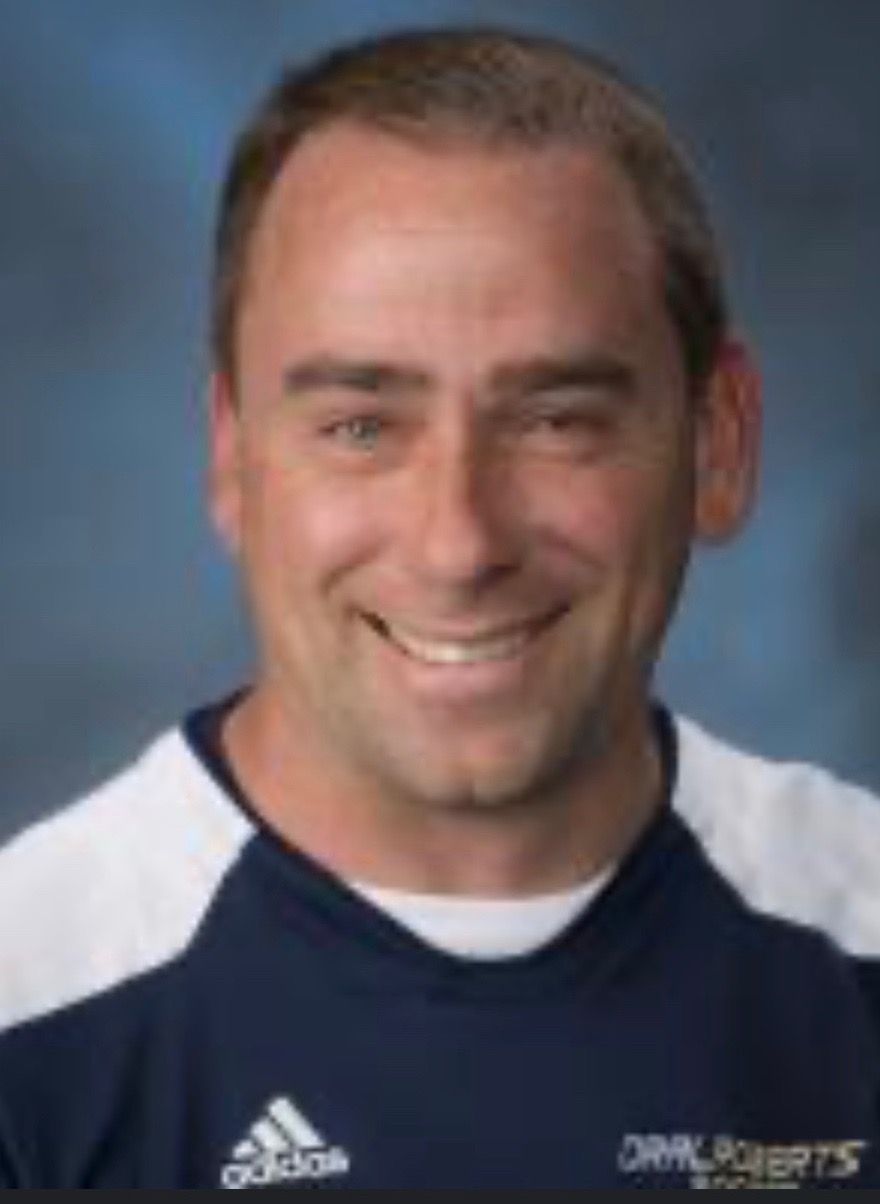 Man smiling, wearing navy shirt with white accents and Adidas logo. Oral Roberts logo visible.