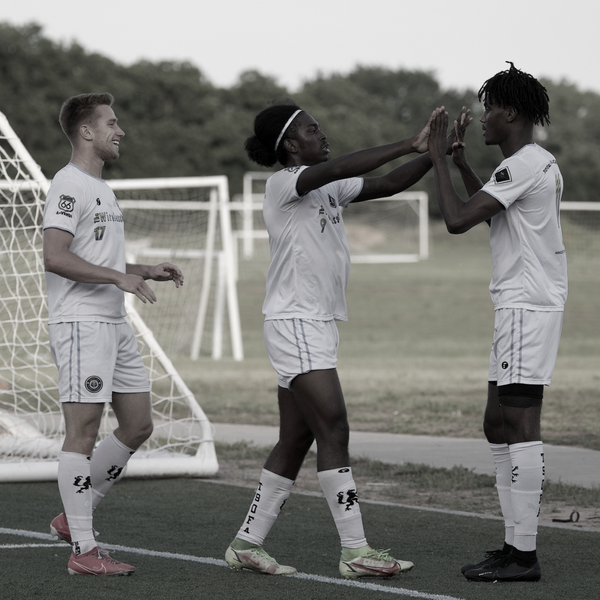 Soccer players celebrate on a green field. Woman high-fives man. Two wear white jerseys, the other looks on.