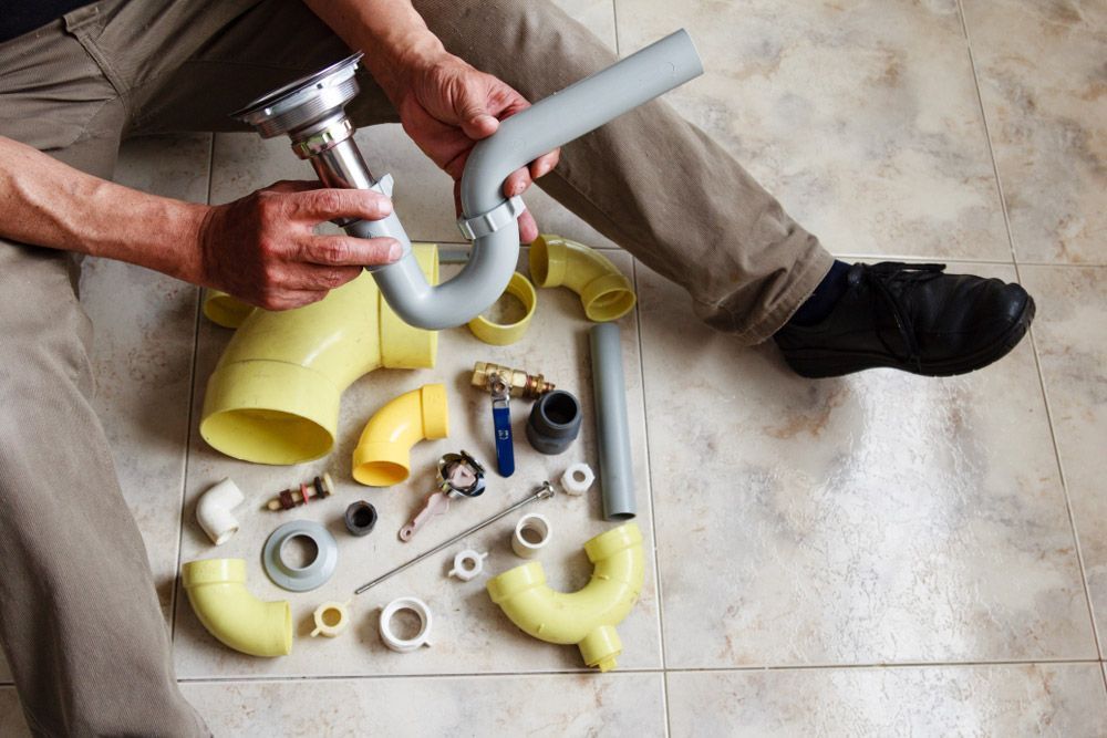 A Man is Sitting on the Floor Fixing a Sink Pipe — All Plumb & Gas in Macksville, NSW