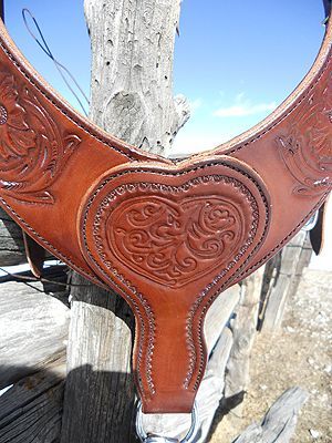 Brown leather horse breast collar with carved heart and floral designs, against a weathered wooden fence.