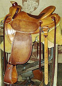 Brown leather Western saddle with stirrups and a cinch, resting on a wooden rack.