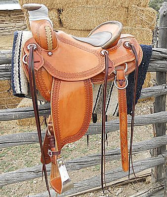 Brown leather western saddle hanging on a wooden fence, with a hay bale in the background.