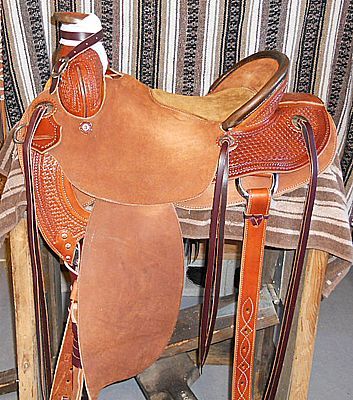 Brown leather Western saddle on a wooden stand. Intricate tooling details, stirrups, and latigo straps.
