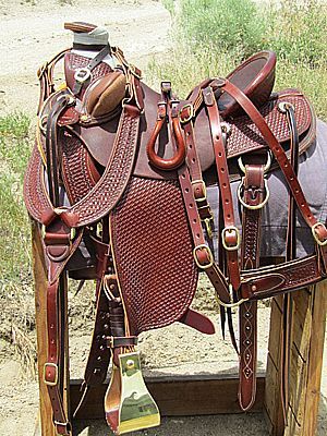 Brown leather western saddle with intricate tooling, on a wooden stand outdoors.